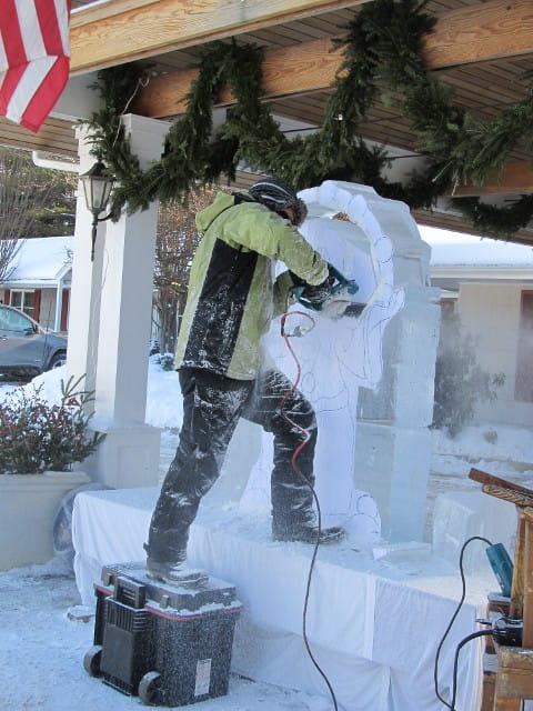 Person in winter gear carving a massive public ice sculpture wall on a sidewalk