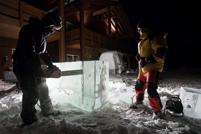 Ice carver finishing a snow structure outside a log cabin at night